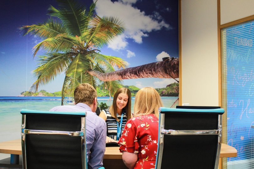 A travel advisor sits behind a desk and talks to two customers.