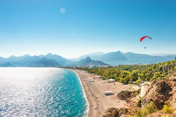 A paraglider soaring over Antalya's beach and mountains, showcasing a stunning coastal landscape and clear blue skies