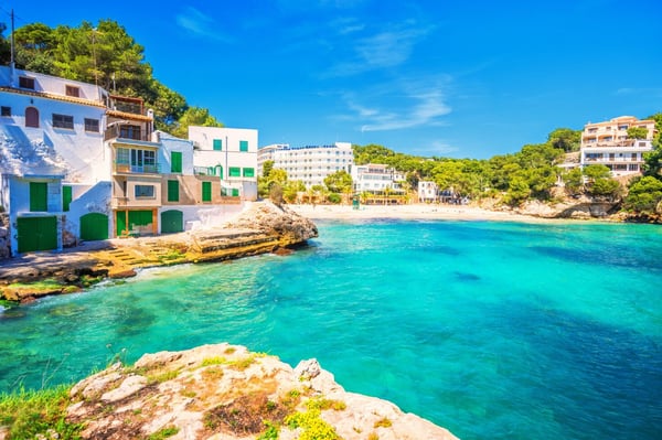 A scenic coastal view of Mallorca, featuring traditional white Mediterranean houses with green shutters along a rocky shoreline. The turquoise waters of a secluded bay gently lap against the shore, with a sandy beach and lush green pine trees in the background under a bright blue sky.