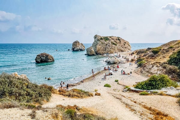 Tranquil Paphos, Cyprus, beach scene with people enjoying a sunny day near turquoise waters and large rocks. Sandy shoreline borders rugged, shrub-covered hills.