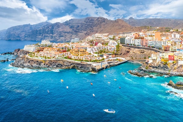 A stunning coastal view of Los Gigantes, Tenerife, showcasing white and pastel-colored buildings perched on rocky cliffs overlooking the vibrant blue Atlantic Ocean, with small boats floating in the water and towering volcanic mountains in the background under a partly cloudy sky.